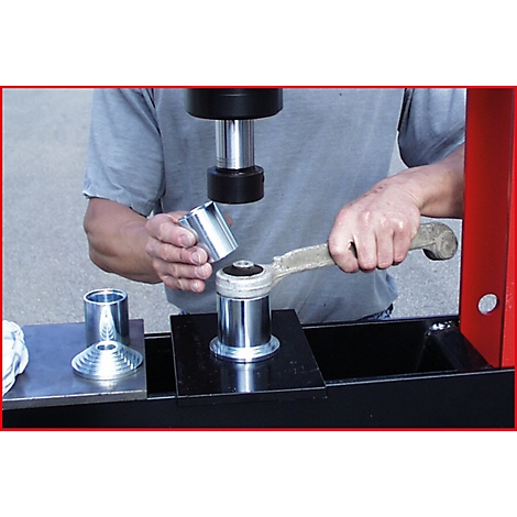 Ein Mann arbeitet mit einer Presse an einem Fahrzeugteil. Er hält einen metallischen Zylinder über ein anderes Teil. Der Hintergrund ist unscharf.